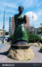 Bronze statue of a historic woman in a dress on a stone base, set in an urban park. Buildings and a flagpole visible in the background.