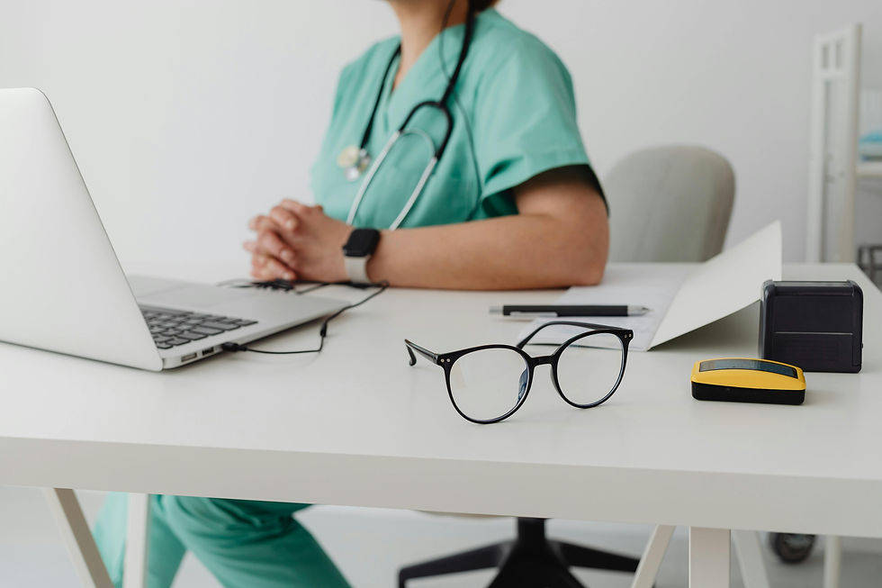 Person in green scrubs at desk with laptop, glasses, and stethoscope. Hands clasped, papers and gadgets nearby in clinical setting.