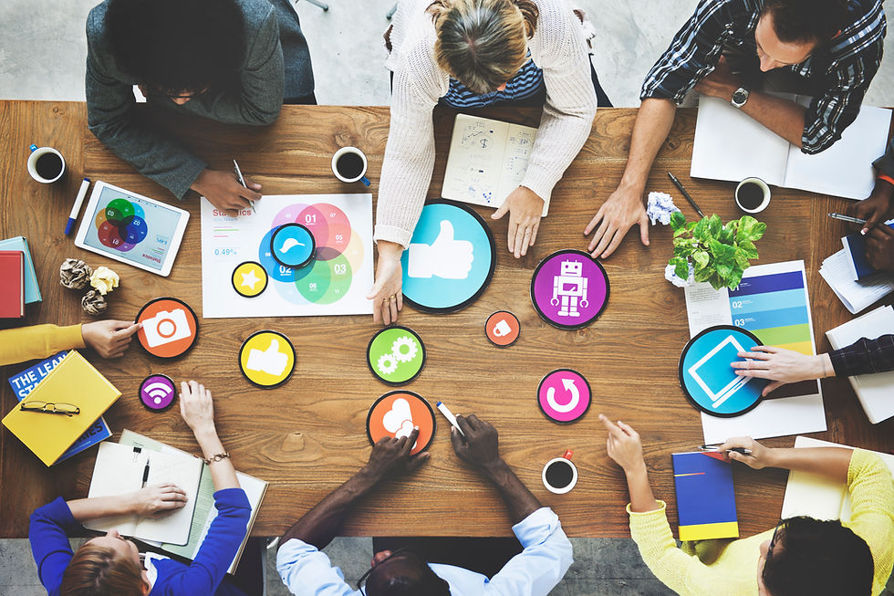 Diverse hands work at a table filled with colorful social media icons, charts, and a plant, embodying teamwork and creativity.