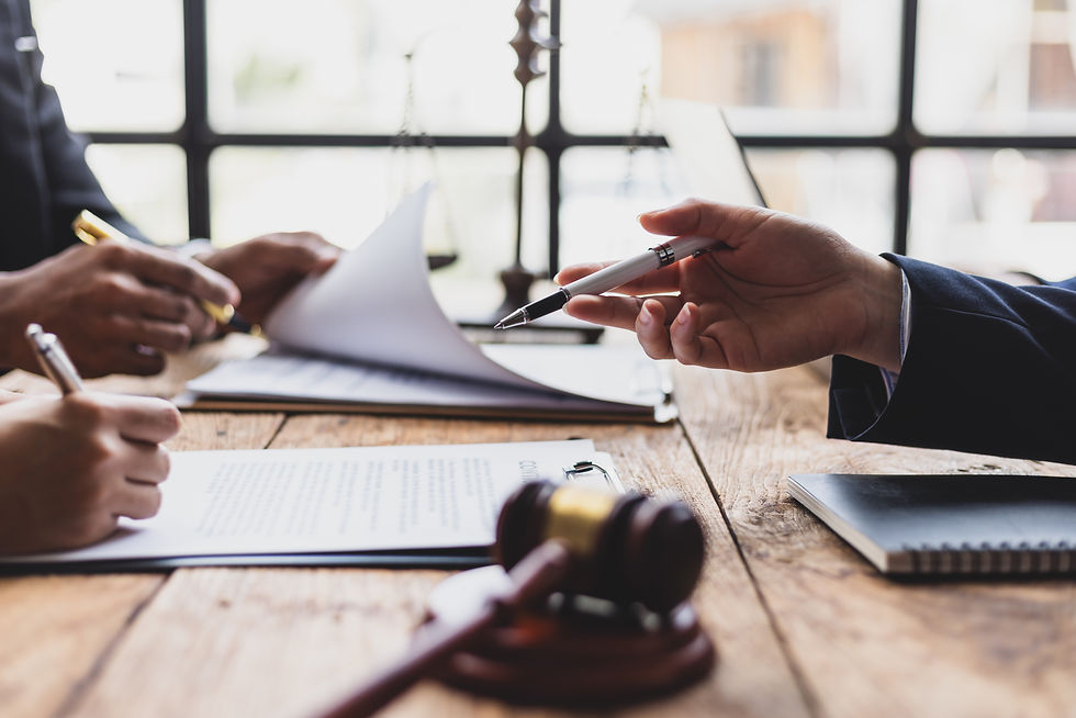Hands hold pens, signing documents on a wooden table. A gavel and legal scales are visible. The setting is a well-lit office.