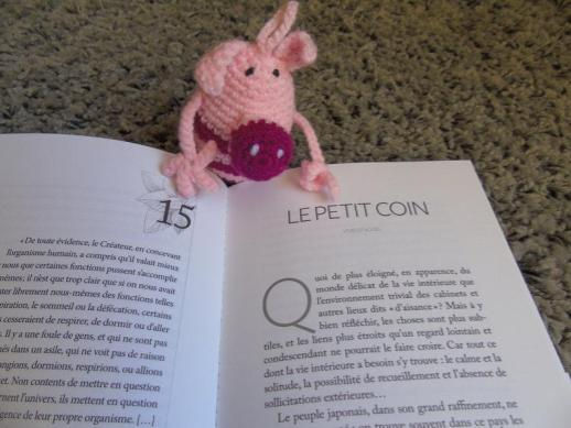 Petit cochon rose tricoté posé sur un livre ouvert, chapitre 15 LE PETIT COIN, sur un tapis.
