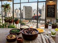 Foto ilustra pratos típicos da feijoada servidos em uma mesa de madeira com a vista da cidade de São Paulo ao fundo