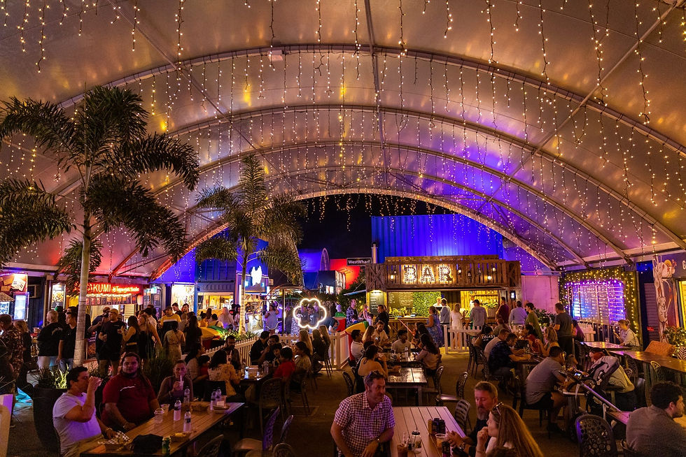 People socialize under a canopy with string lights. A bar and food stalls are in the background. The setting is colorful and lively.