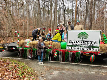 Eleven people bundled in winter clothes sit on hay bales on a trailer decorated as a parade float with red, green, and silver Christmas tinsel and a large corrugated plastic sign that reads Garrett's Tree Service LLC Stump Grinding 919-475-7804.