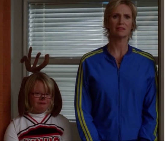 Becky the Rein-deer and Sue Sylvester from Fox's Glee. A young cheerleader wearing reindeer antlers and a painted black nose stands beside an adult in a blue track jacket; both face forward indoors before window blinds, looking serious.