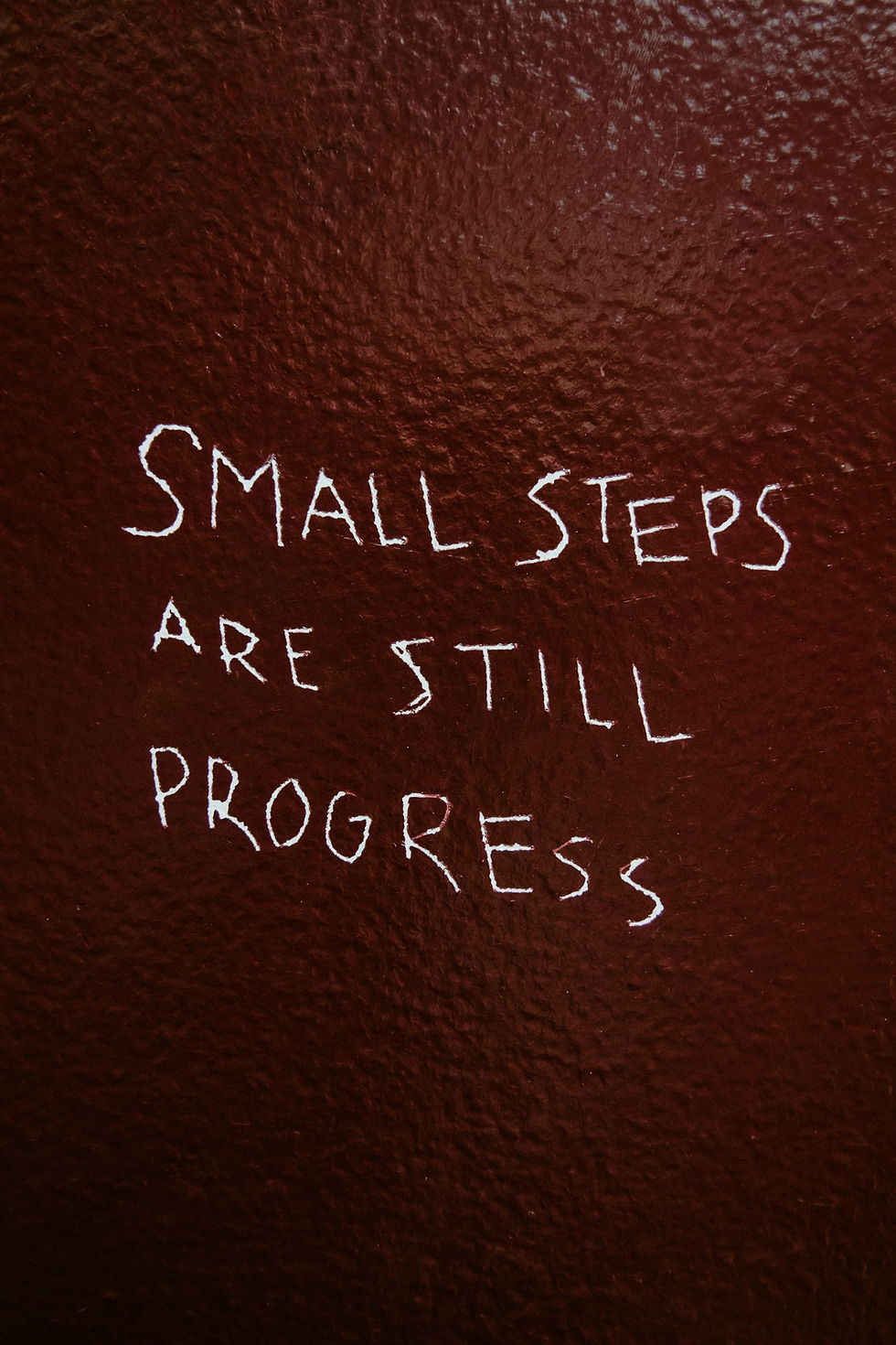 White text on a dark red wall reads "Small steps are still progress," conveying motivation. The wall texture is rough.