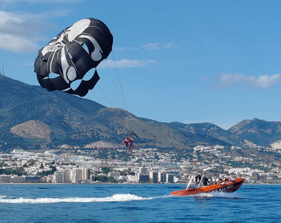 Two people parasail over the ocean, pulled by an orange boat with "PARASAILING" on the side. Mountains and cityscape in the background.