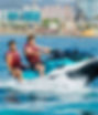 Two men in red life jackets ride a teal jet ski with 6 on the side across blue water near a city waterfront, splashing hard.
