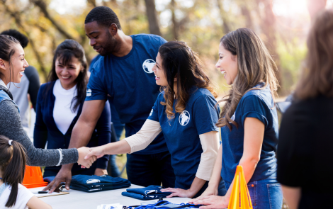 Volunteers shaking hands at an outdoor event, smiling. People and trees in background.
