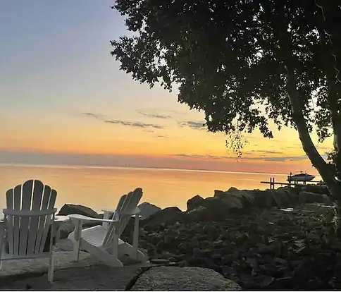Adirondack chairs facing Lake Erie at sunset under the trees near The Lakeside House, with soft orange light over calm water