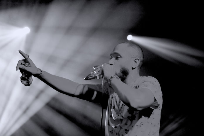 Mos Def in concert holding a mic in his left hand, with his right hand stretched to the crowd