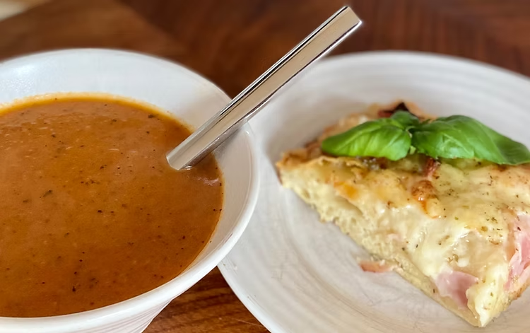 Skål med tomatsoppa och sked bredvid en bit focaccia med basilika på vit tallrik, på ett träbord.