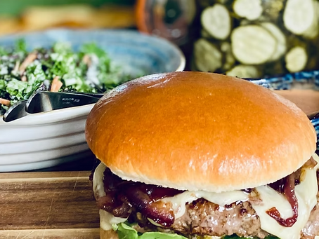 Stor hamburgare på skärbräda med salladsskål och inlagda gurkor i bakgrunden.