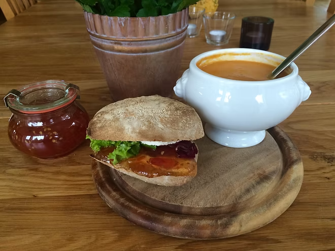 Smörgås med sallad och pålägg på träbricka, bredvid soppa i vit skål och en burk tomatmarmelad på träbord.