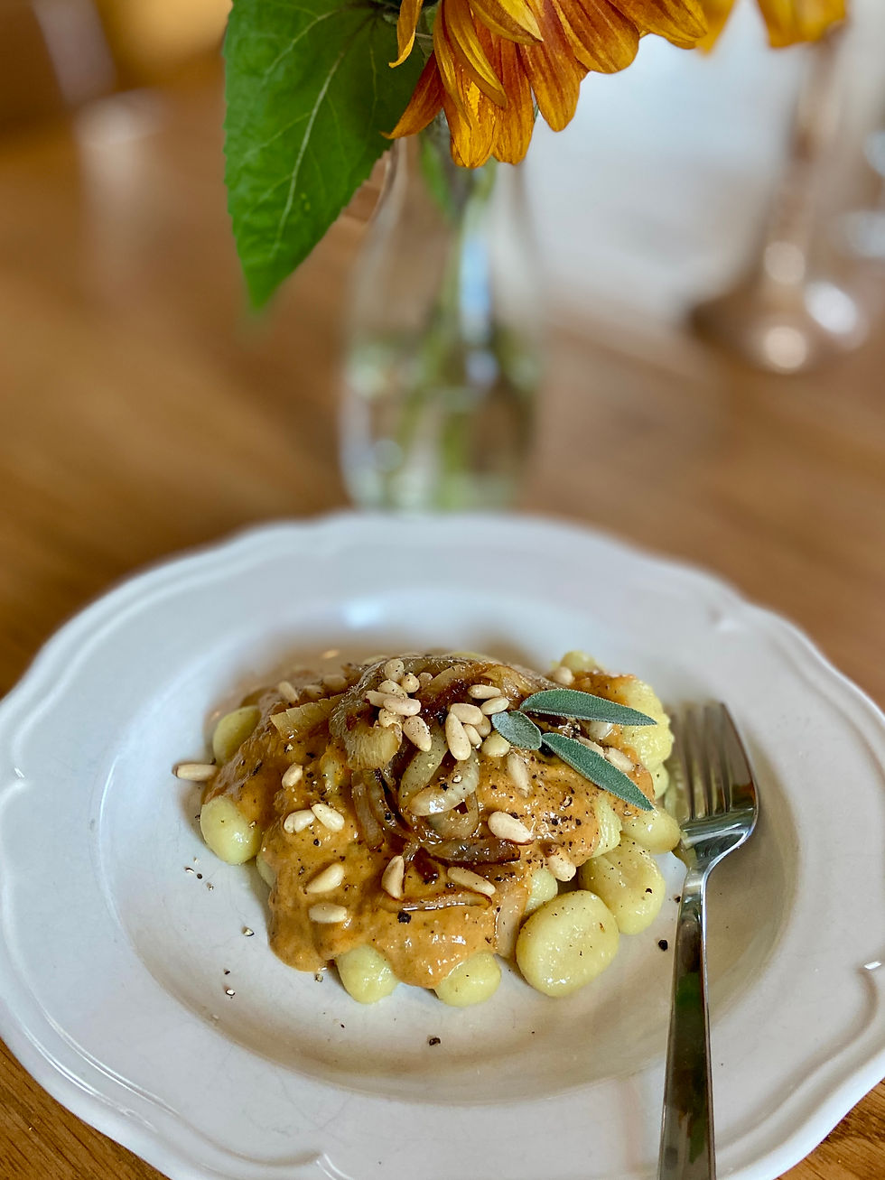 Tallrik med gnocchi, pumpasås, stekt lök, pinjenötter och salviablad, med gaffel på ett bord och suddig blomma i bakgrunden.