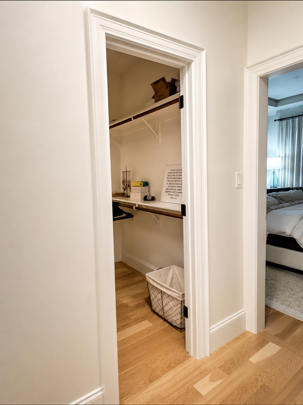 Doorless walk-in closet mid-makeover with white walls, wood and white shelving, a wire laundry basket, and a framed sign — the master bedroom visible through the doorway beside it."
