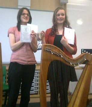 two young ladies holding a trophy and certificates behind a small harp