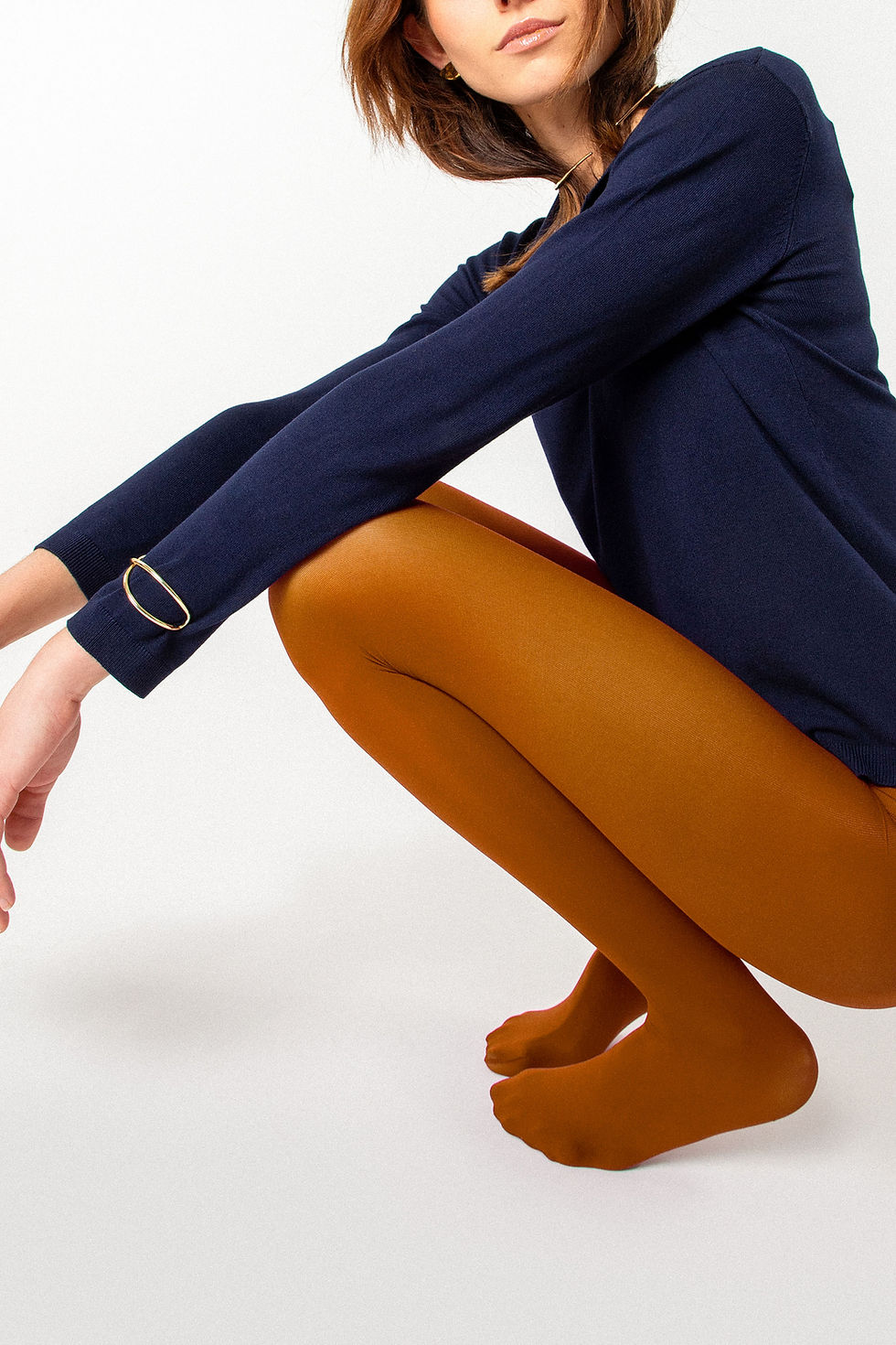 Woman in blue sweater and orange leggings squats on a white background, wearing gold jewelry. Neutral expression, minimalist setting.