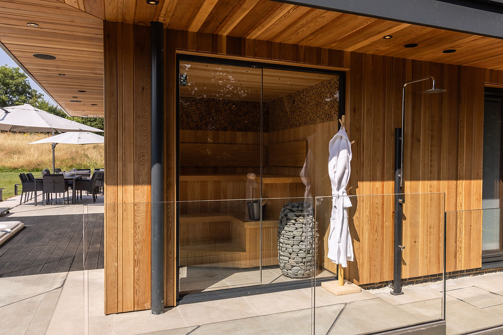 Modern outdoor sauna with glass doors, wooden panels, and stones. A white robe hangs nearby. Patio furniture and lawn in the background.