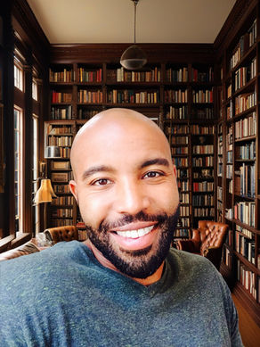 A smiling man in a grey sweater, in a library filled with books.