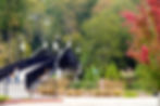 Mother and child walk across a black bridge in a park. Background features green and red foliage, lampposts, and hanging banners.