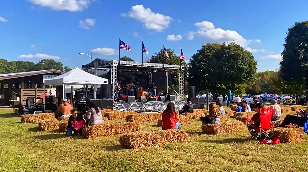 Band performing on outdoor stage with guitar, bass, banjo, and mic. Audience in folding chairs, fall flowers and wooden building in background.