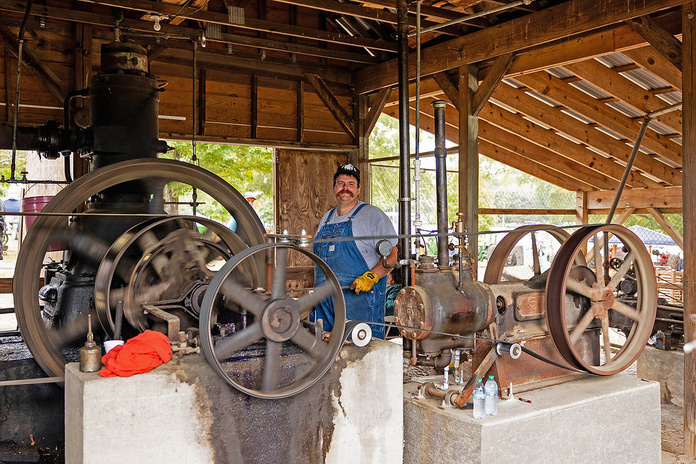 Man wearing overalls and hat stands by a vintage engine at Cotton Ginning Days