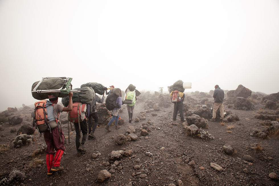 Hikers carry heavy loads up a rocky, misty mountain trail. The scene is overcast, with muted colors, conveying a mood of determination.
