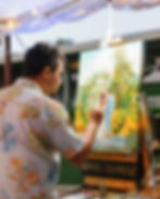 Wedding Artist paints a colorful wedding scene outdoors, wearing a floral shirt. Easel bears the name "ARIEL QUIROZ." Soft evening light.