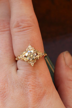 Round lab grown diamond filigree antique style engagement ring with small accent diamonds, set in yellow gold, worn on a finger.