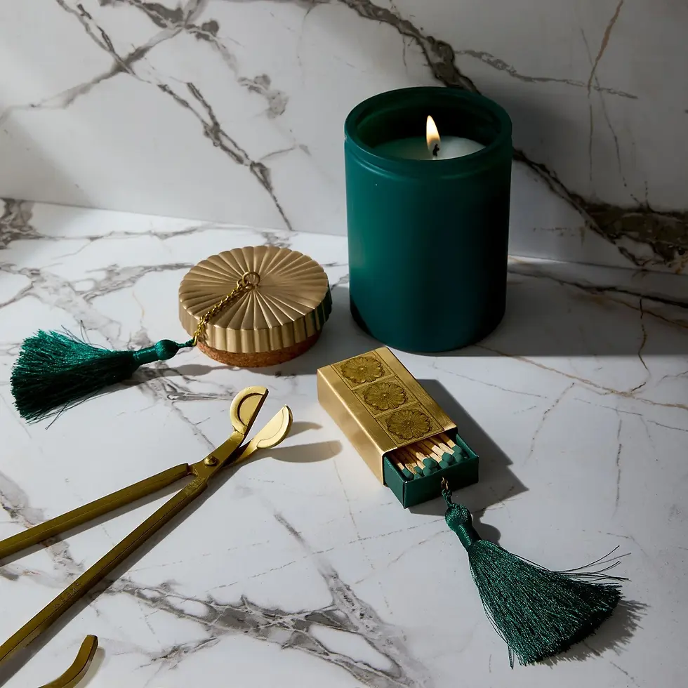 A green candle, gold wick trimmer and brass matchbox with dark green tassel on a white and black marble countertop.