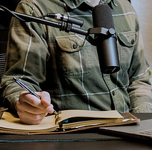 Person with a green flannel talking into a podcast microphone and writing in a notebook