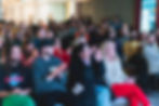 Audience clapping in a brightly lit room, smiling and engaged. Diverse group, colorful clothing, text on screen in the background. Mood: joyful.