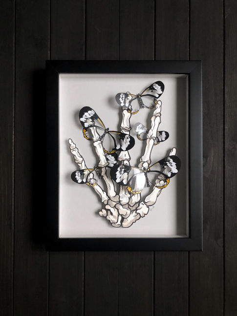 Dimensional contemporary paper art of a human skeleton hand surrounded by transparent glasswing butterflies made of paper, and titled "Fragile"