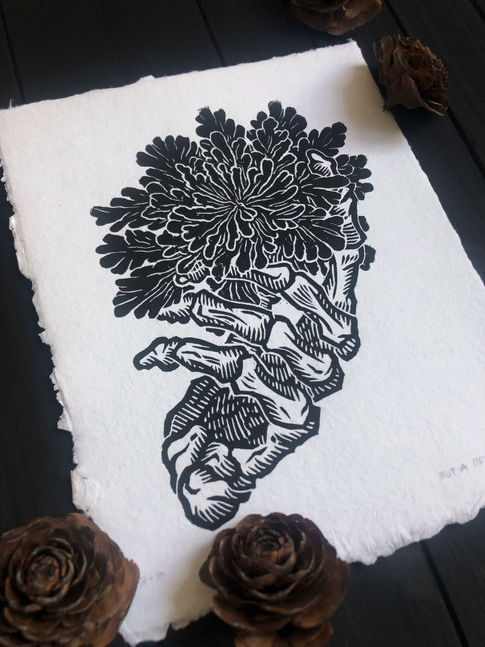Linocut print of a skeleton hand holding an opening resurrection fern, printed on handmade recycled paper, and titled "But A Dry Spell"