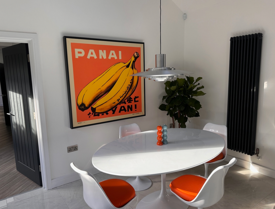 Photo of a completed design in a new build property. The photo illustrates design classics. A louis poulsen pendant light hangs above Eero Saarinen tulip dining chairs with an orange seat and oval shaped white dining table. On the wall is a large, bold graphic design print with an orange back drop, bananas and the word Panai at the top in an Andy Warhol-esque style. 