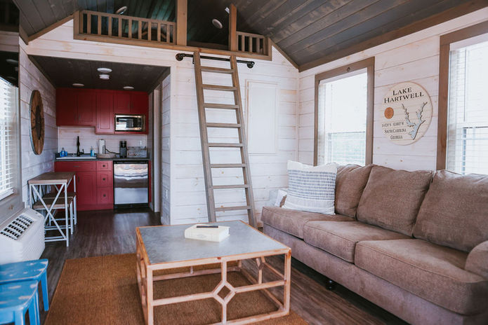 Living room in tiny cabin with beige couch and grey/wood farmhouse coffee table. Access to overhead lofts.