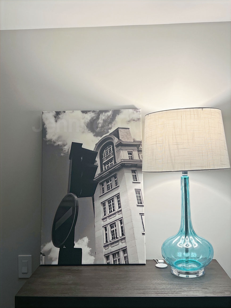 A black and white canvas photo of a Paris building and traffic light placed on top of a dresser with a blue lamp to the right.