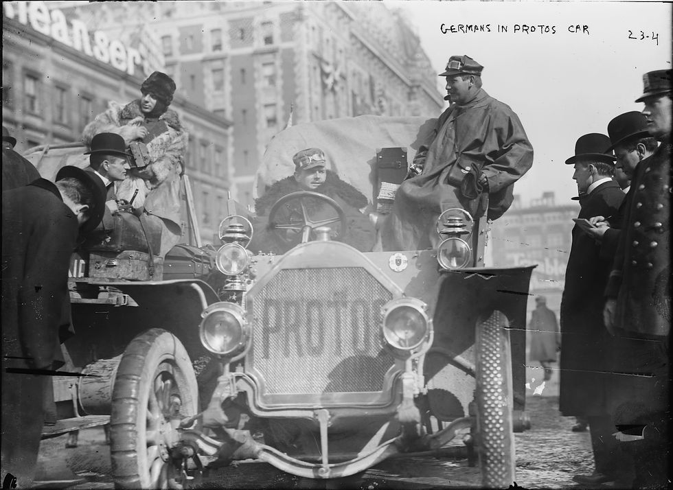 Startaufstellung des New-York–Paris-Rennens 1908: mehrere offene Autos mit Schildern „New York–Paris“, dicht besetzte Fahrzeuge mit warm gekleideten Fahrern und Beifahrern, Zuschauer stehen am Straßenrand und schauen aus den Fenstern der Häuser im Hintergrund.