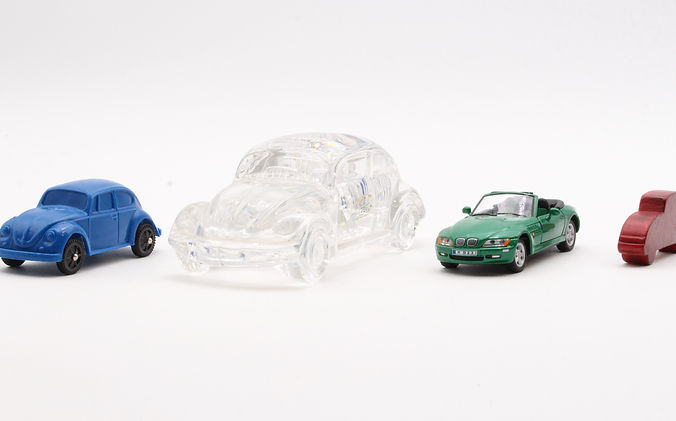 Vier unterschiedliche Miniaturautos nebeneinander vor weißem Hintergrund: links ein blaues Kunststoffauto im Käfer-Stil, daneben ein größeres durchsichtiges Käfermodell aus Glas oder Acryl, rechts ein grünes detailreiches Cabrio-Modellauto und ganz rechts ein schlichtes, rotes Auto-Symbol aus Holz.