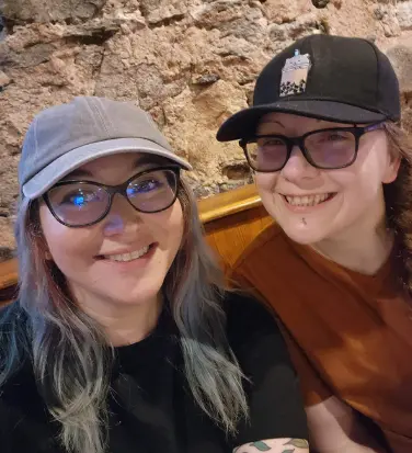 Two people wearing glasses and baseball caps smile in front of a stone wall. One wears a black shirt, the other a brown shirt.