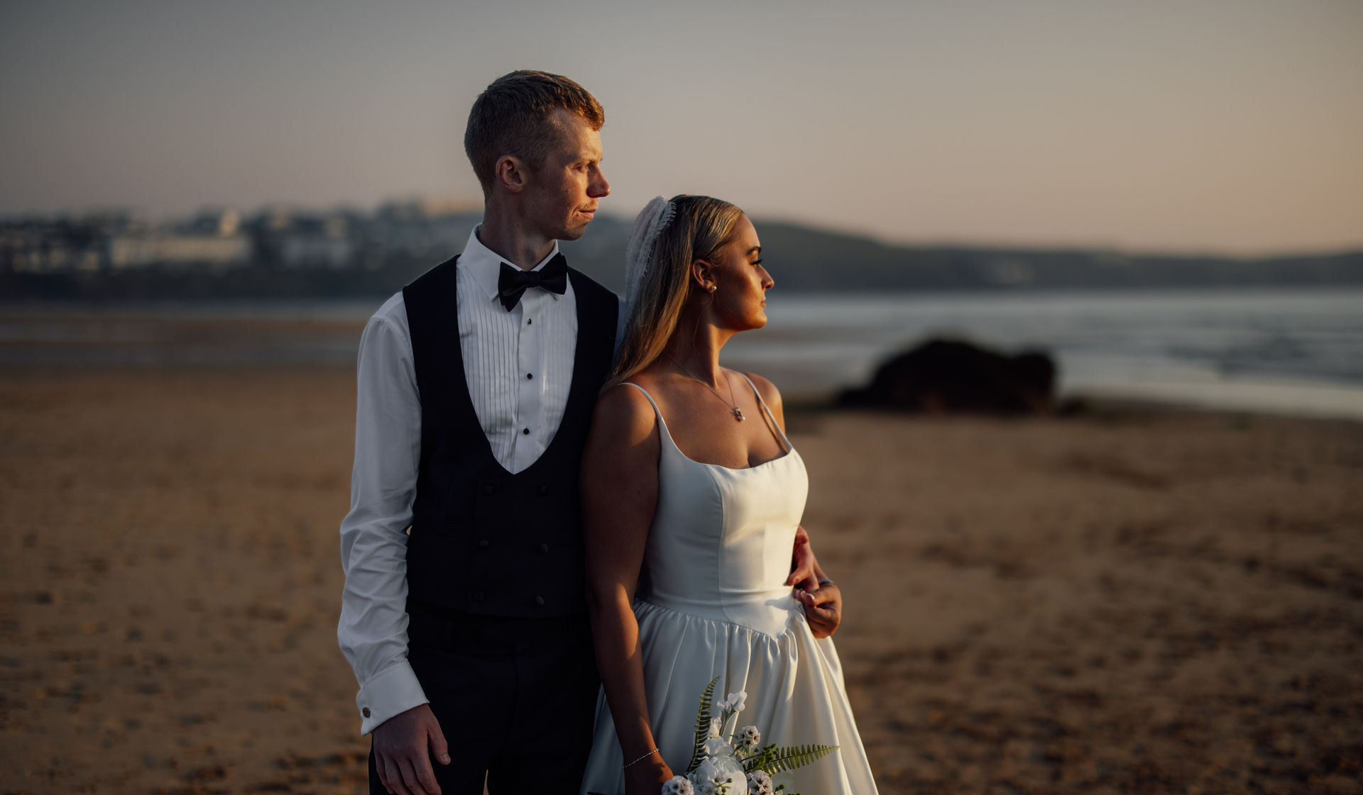 A bride and groom stand on a beach in Devon at sunset after their wedding. Captured by award winning, documentary photographers, Fox and Co Photography.