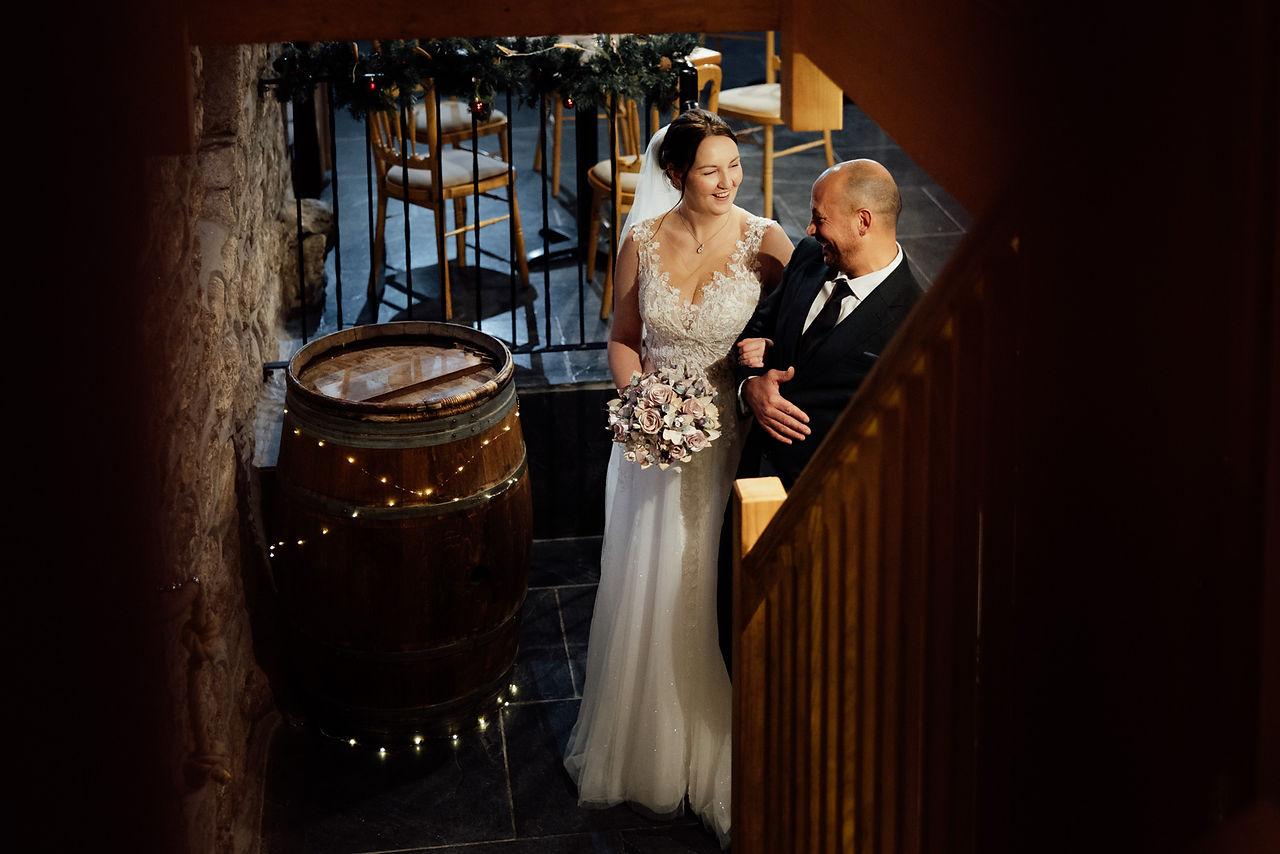 The bride and groom wait at the bottom of stairs at Knightor Winery smiling at each other.
