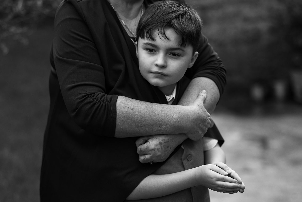 A child waits patiently whilst his mother hugs him from behind
