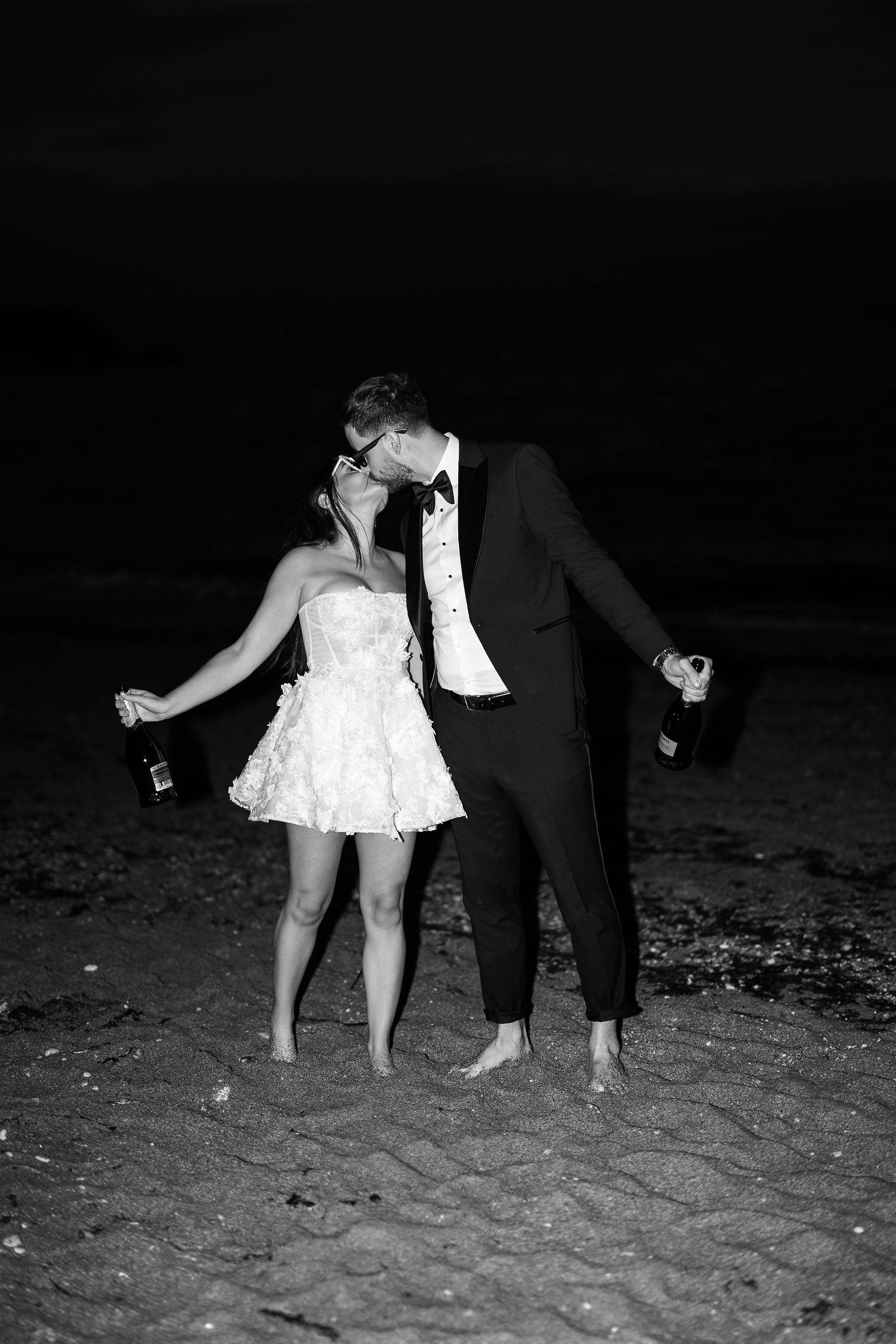 An editorial image of a bride and groom sharing a kiss whilst holding a bottle of champagne. 