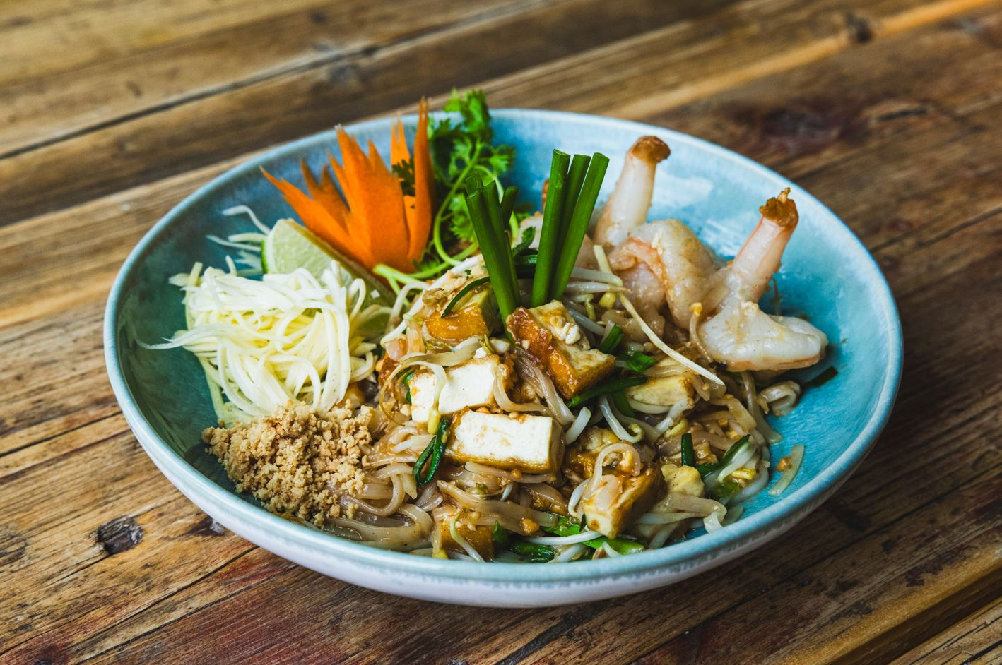 Pad Thai mit Shrimps und Gemüse in einer blauen Schüssel auf Holzuntergrund.