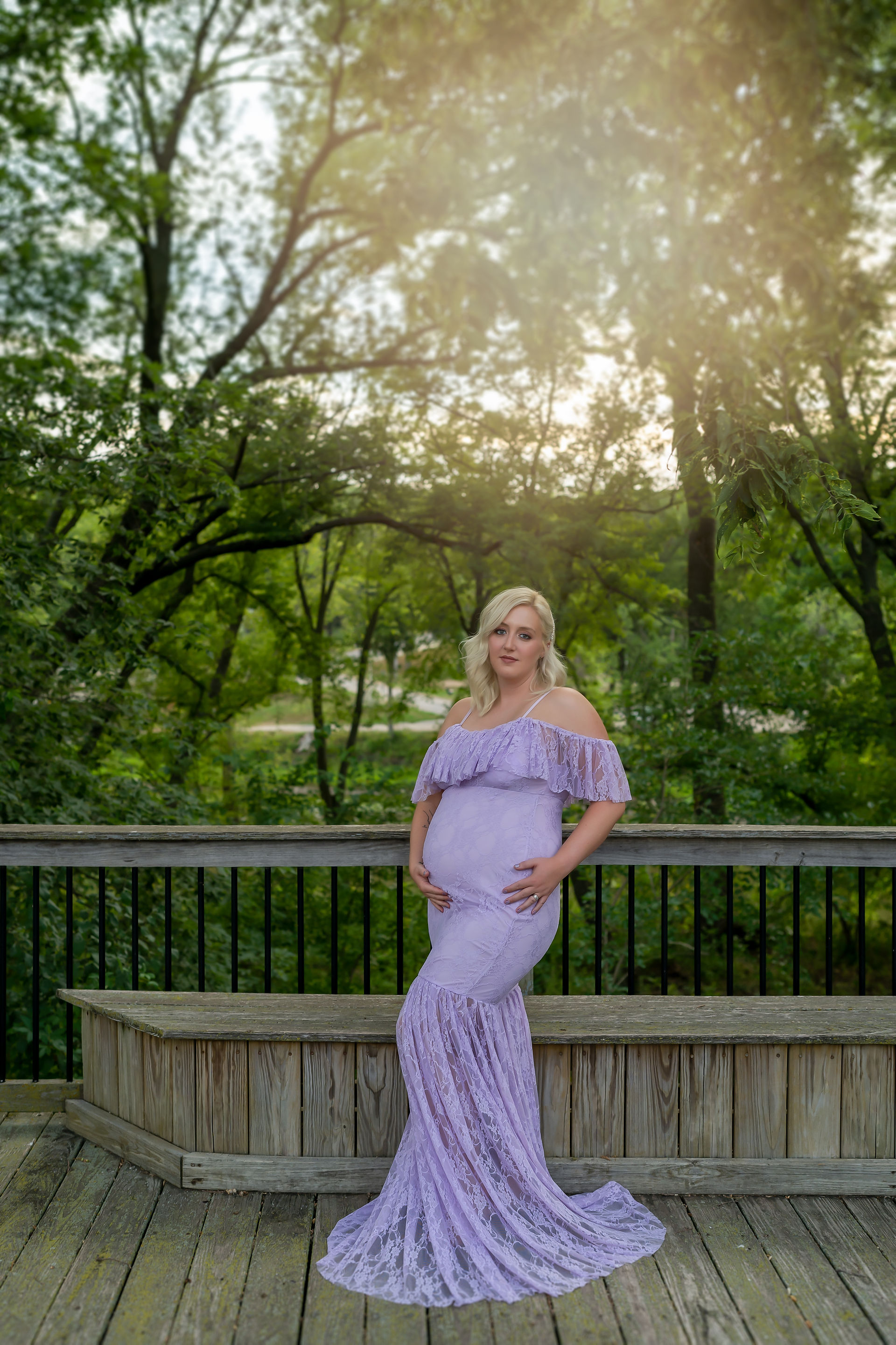 Des Moines and Norwalk, Iowa family photographer - pregnant blonde female wearing a light purple gown is posed standing on a wooden platform with trees and the Des Moines River in the background.