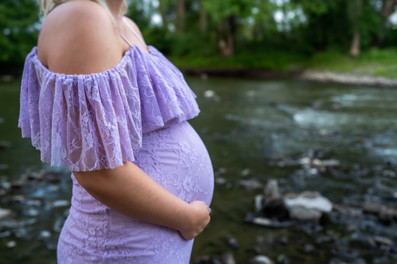 Des Moines and Norwalk, Iowa family photographer - pregnant blonde female wearing a light purple gown is posed standing in front of the Des Moines River with a close up shot of her belly.