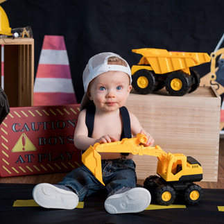 Des Moines, Iowa photographer - infant boy is sitting in studio for a constuction themed photo session. He is wearing denim jeans, suspenders, and backwards cap and is holding a large toy truck..