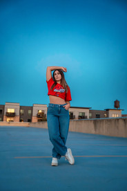 Des Moines, Iowa senior photography - teenage girl with dark hair and a cropped Iowa State Cyclone shirt poses on the rooftop during blue hour with her hand over her head and feet crossed at the ankles.
